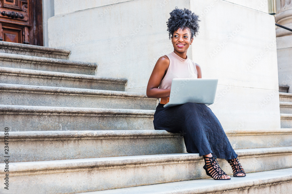 Way to Success. Young African American woman with afro hair, wearing sleeveless top, black skirt, sandals, sitting on stairs outside office building in New York, working on laptop computer, smiling..