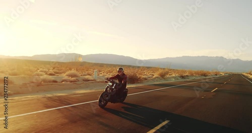 Happy man riding vintage black motorcycle down desert highway at sunset 