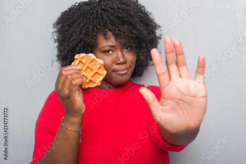 Young african american woman over grey grunge wall eating belgium waffle with open hand doing stop sign with serious and confident expression, defense gesture