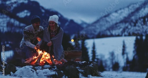 Couple Sitting by the Burning Campfire With Winter Mountains in Background. SLOW MOTION. Fairytale snow covered view with fire burning outdoors at dusk. 