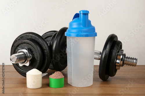 Empty protein shake sport bottle, scoops with powders and dumbbells on table