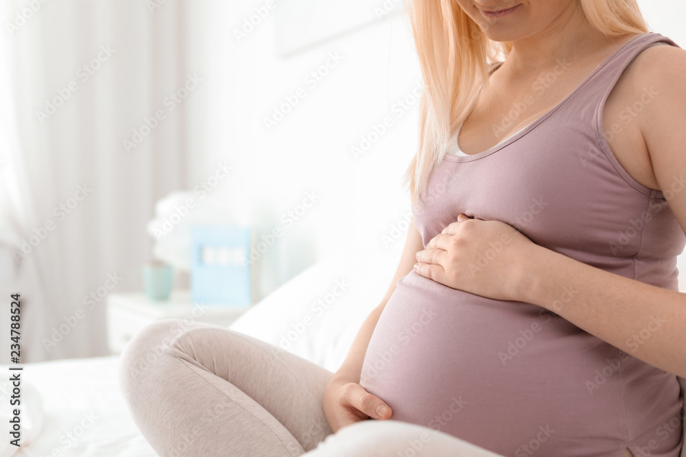 Pregnant woman sitting on bed in light room. Space for text Pregnant woman sitting on bed in light room. Space for text