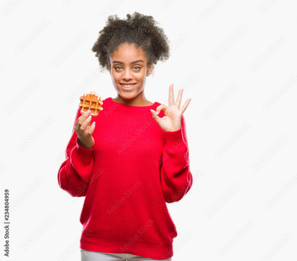 Young beautiful afro american woman eating waffle over isolated background doing ok sign with fingers, excellent symbol Young beautiful afro american woman eating waffle over isolated background doing ok sign with fingers, excellent symbol