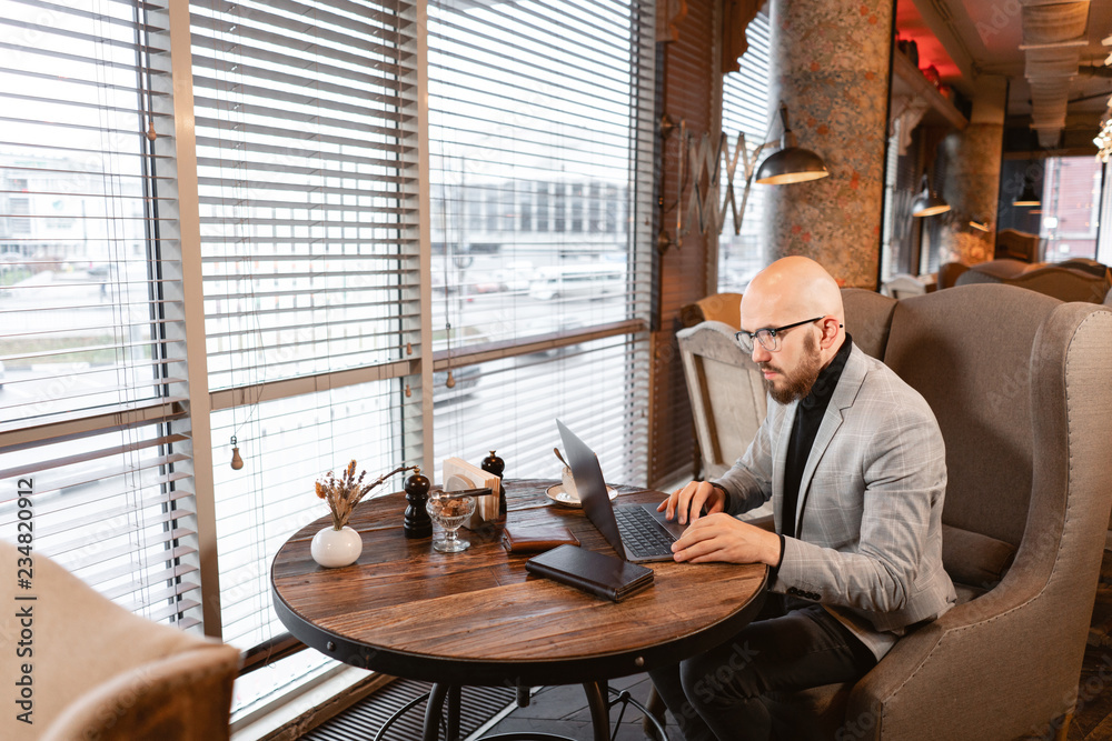 Portrait of young man with beard in the glasses looking to the monitor laptop. Successful people, businessman in comfortable cafe . Reading text during work on netbook Portrait of young man with beard in the glasses looking to the monitor laptop. Successful people, businessman in comfortable cafe . Reading text during work on netbook