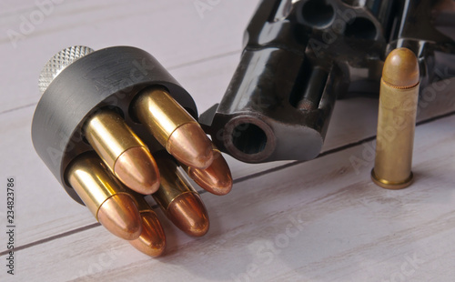 A loaded speed loader loaded with 38 special full metal jacket bullets in front of a black revolver with a single bullet next it  on a white wooden background