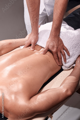 two young man, 20-29 years old, sports physiotherapy indoors in studio, photo shoot. Physiotherapist massaging patient lower back with his hands close-up.