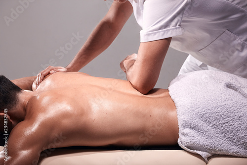 side view, two young man, 20-29 years old, sports physiotherapy indoors in studio, photo shoot. Physiotherapist massaging muscular patient lower back with his hands.