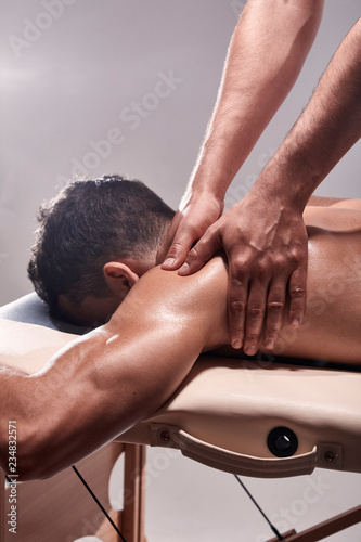 side view, two young man, 20-29 years old, sports physiotherapy indoors in studio, photo shoot. Physiotherapist massaging patient shoulder with his hands close-up.