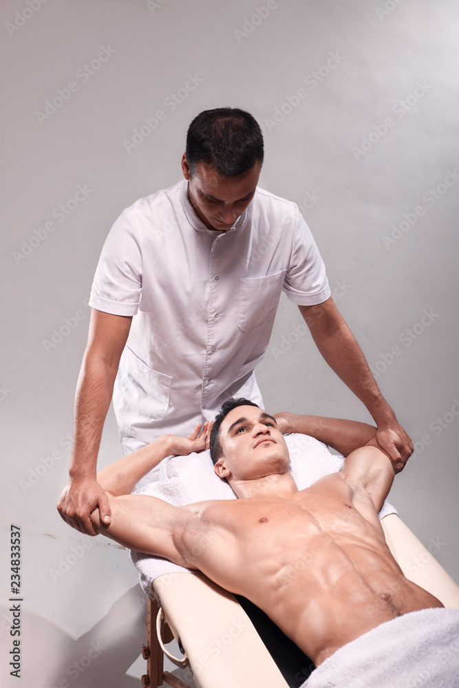 Obraz premium two young man, 20-29 years old, sports physiotherapy indoors in studio, photo shoot. Physiotherapist stretching muscular patient arms with his hands.