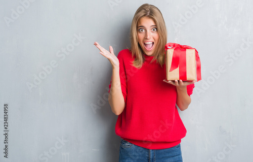 Beautiful young woman over grunge grey wall holding in love a present very happy and excited, winner expression celebrating victory screaming with big smile and raised hands