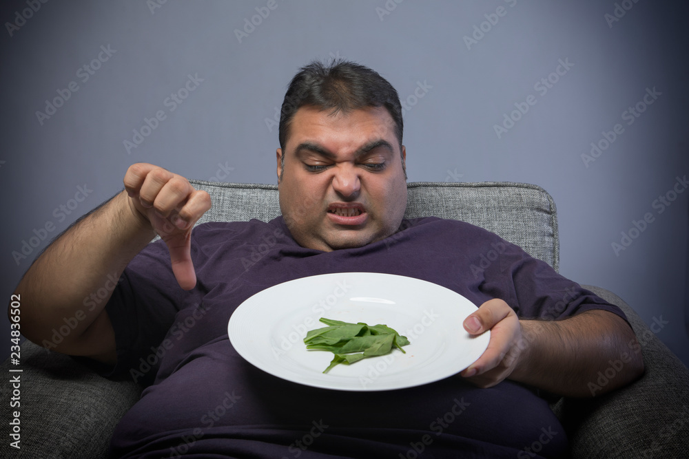 Unhappy obese man holding a plate with few spinach leaves and showing a thumbs down sign Unhappy obese man holding a plate with few spinach leaves and showing a thumbs down sign