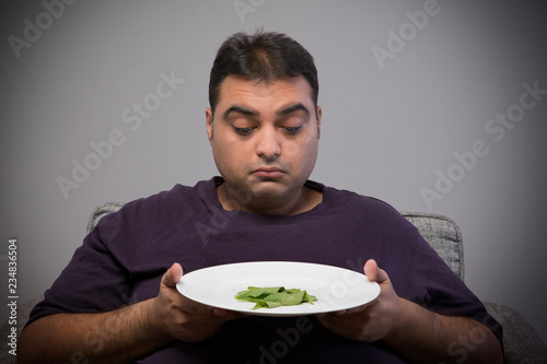 Obese man holding a plate with few spinach leaves and looking at it sadly