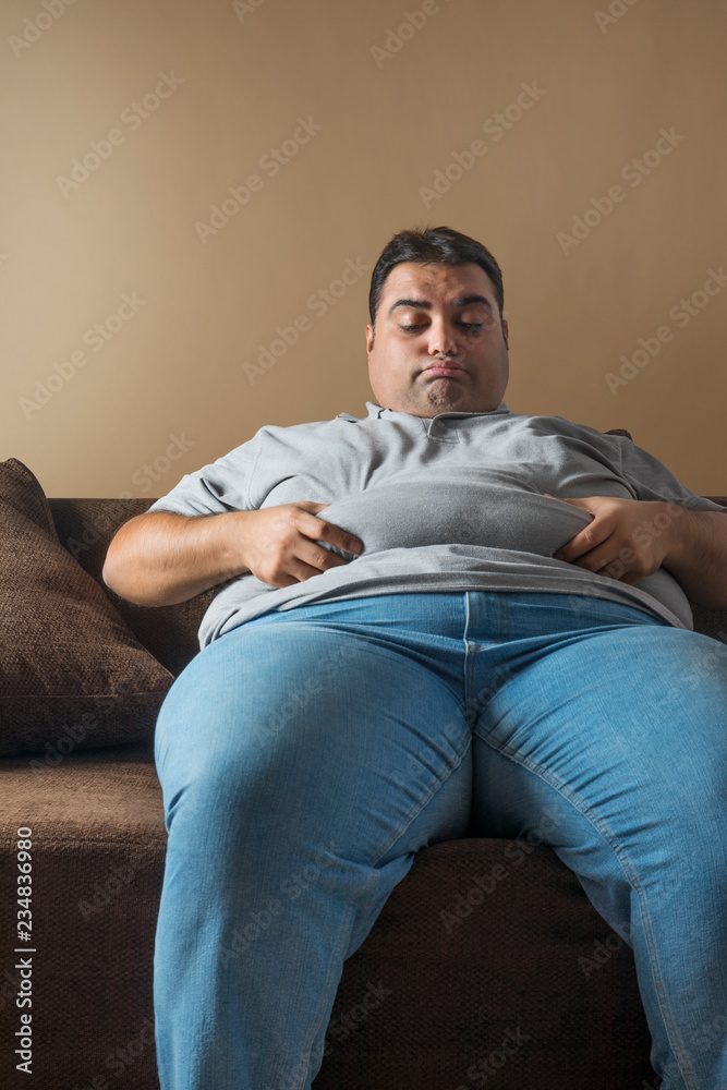Unhappy obese man sitting on sofa holding his belly fat with both hands  Stock Photo | Adobe Stock