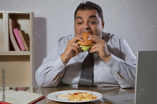 Overweight man eating burger sitting at his desk in office with laptop in front