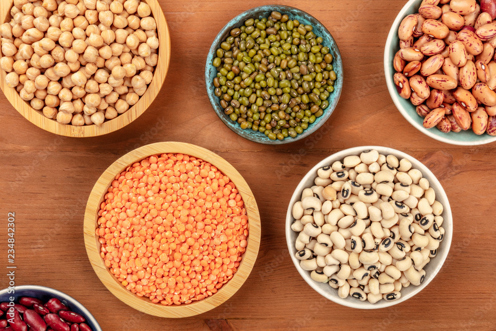 A photo of various types of legumes, shot from the top on a dark rustic wooden background. Different beans, lentils, chickpeas, soybeans