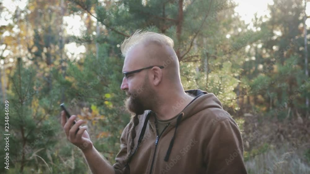 Irritated guy with glasses and a beard shouting and swearing obscenely on the phone, holding the phone in his hand against the background of the autumn park Irritated guy with glasses and a beard shouting and swearing obscenely on the phone, holding the phone in his hand against the background of the autumn park