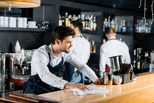 adult smiling barman cleaning wooden counter with cloth