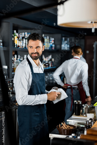 smiling handsome bartender polishing glass near counter