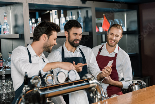 handsome adult barmen smiling and using smartphone at workplace