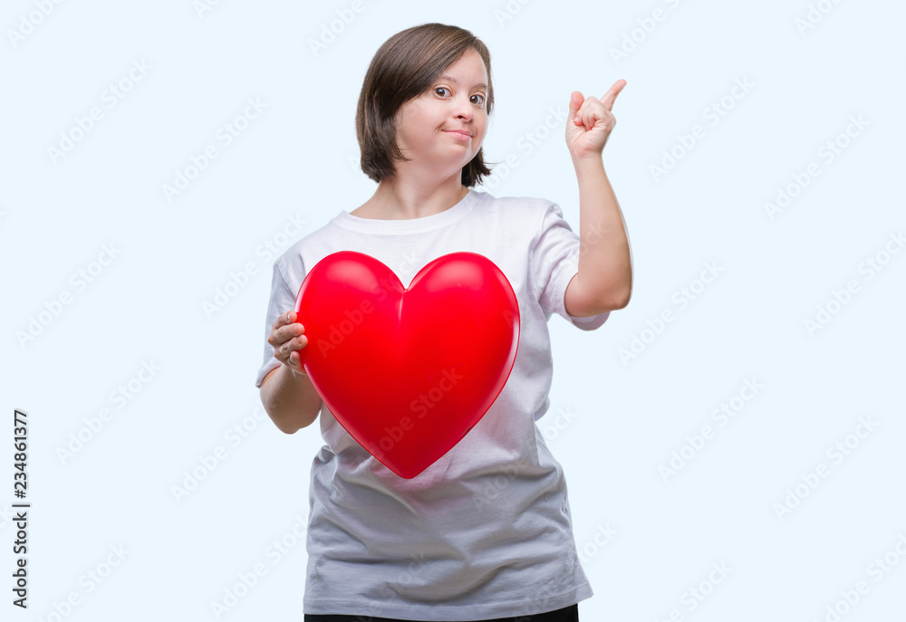 Young adult woman with down syndrome holding red heart over isolated background very happy pointing with hand and finger to the side Young adult woman with down syndrome holding red heart over isolated background very happy pointing with hand and finger to the side