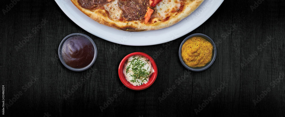 A kitchen dark wooden desk with food standing on it: delicious pizza on white plate and three small round bowls full of sauces, all arranged in nice looking composition.
