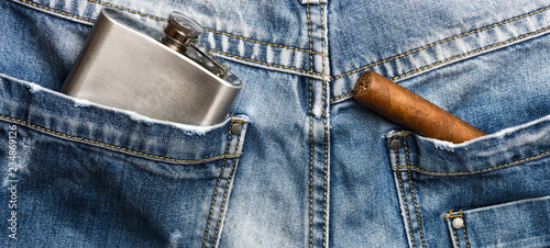 Cigar and alcohol drink in back pocket of pants. Elite alcohol and cigar. Gourmet concept. Habit to smoke elite tobacco. Pocket of denim pants staffed with cigar and metal flask denim background