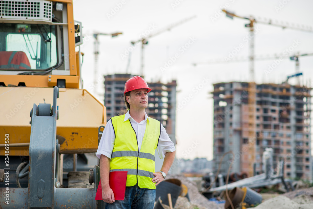 Engineer in front of excavator on a construction site with buildings and cranes in the background Engineer in front of excavator on a construction site with buildings and cranes in the background