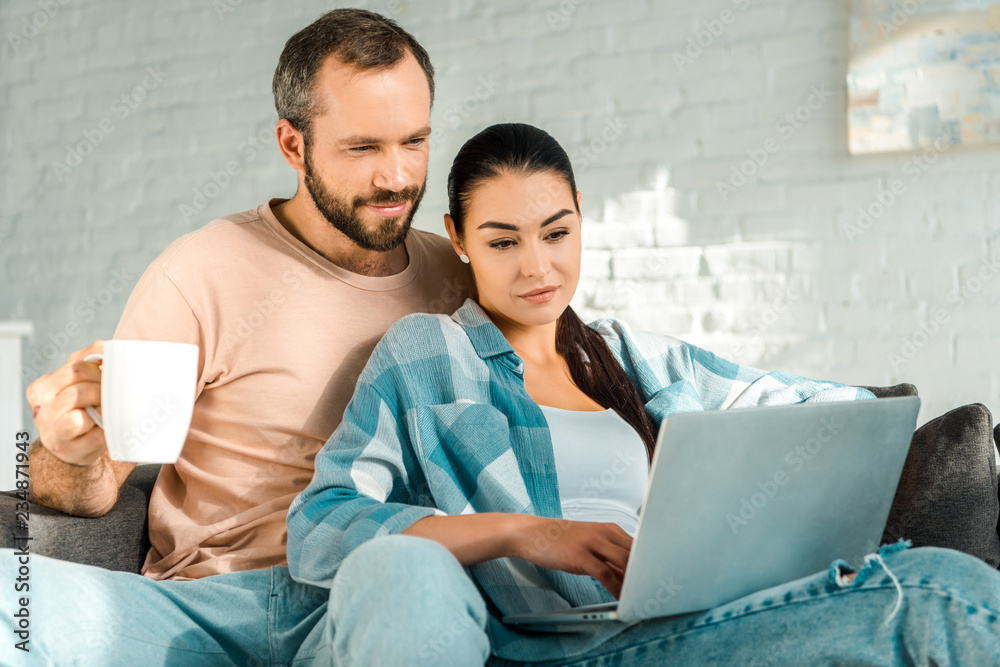 Fototapeta premium handsome husband holding cup of coffee while wife using laptop on couch