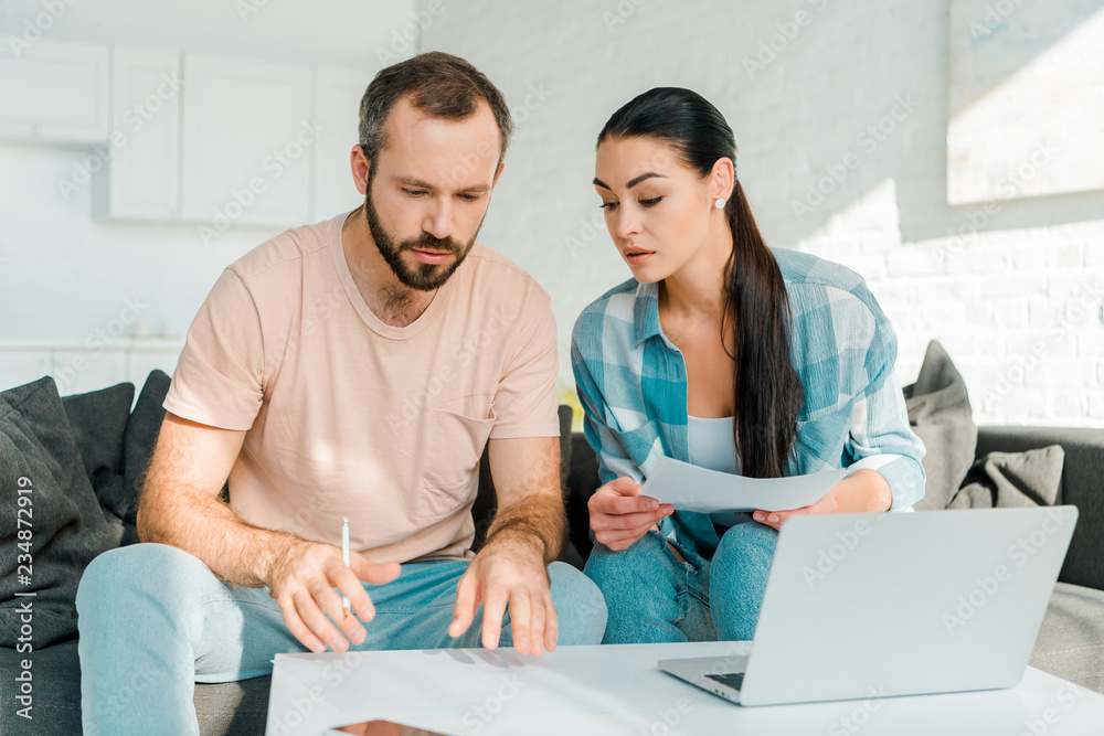 concentrated couple sitting on couch, filling papers, using laptop and planning concentrated couple sitting on couch, filling papers, using laptop and planning