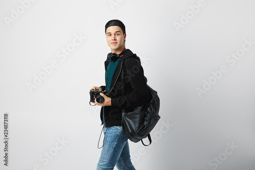 Smiling guy with a black backpack on his shoulder dressed in a dark green t-shirt, jeans, sweatshirt and a cap holds a camera in his hand on a white background