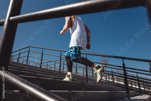 Athletic man with headband on his head dressed in the white t-shirt, black leggings and blue shorts is running up the stairs outside on a sunny day