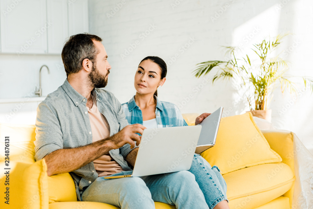 handsome husband using laptop while beautiful wife holding notebook and sitting on couch