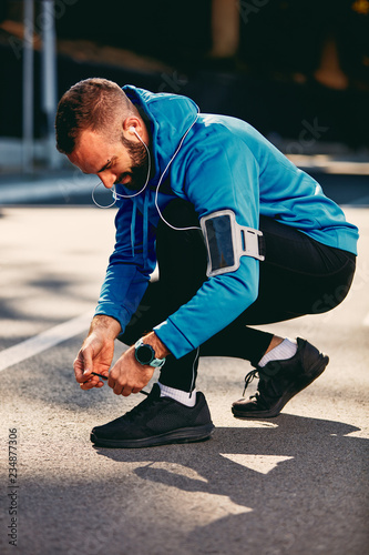 Male runner crouching and tying shoelace. Sportswear on, earphones in ears. Healthy lifestyle concept.
