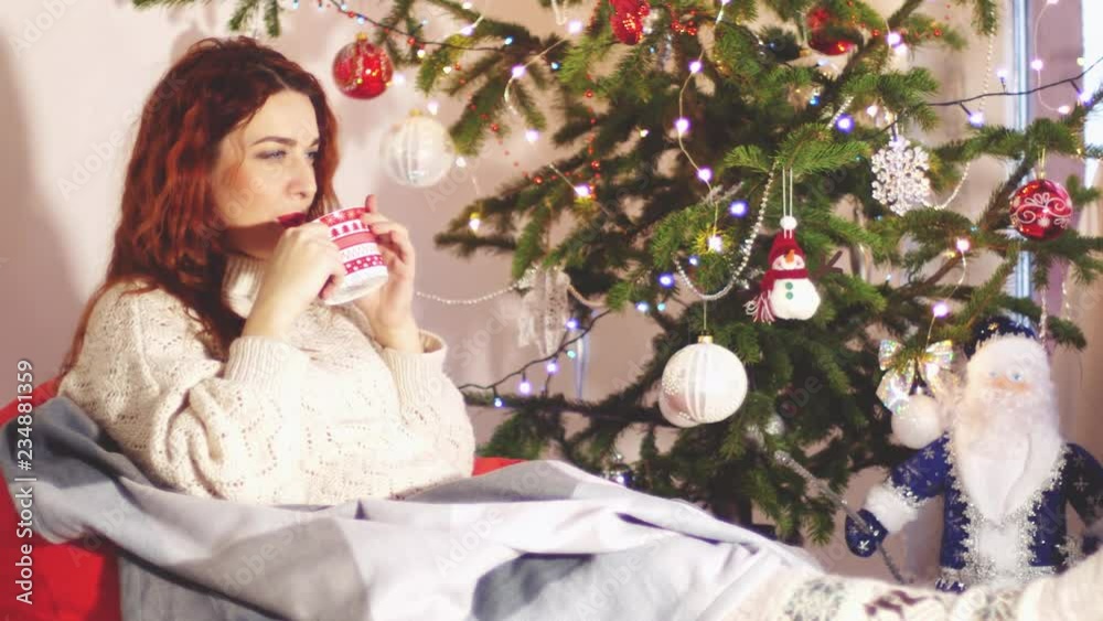 Portrait of beautiful young woman wearing stylish christmas sweater and socks sitting on comfortable sofa and drinks tea by christmas fir-tree. 3840x2160