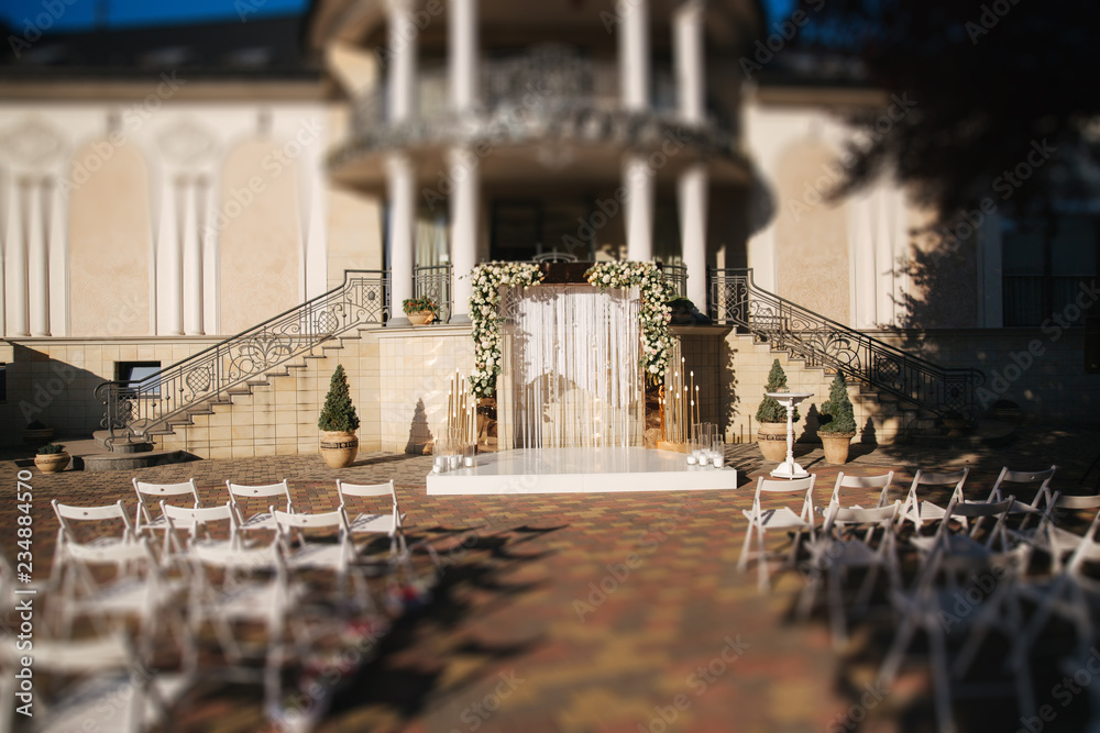 Weddind decoration on open air. Floral decor of a beautiful white and gold arch. Beautiful beckground view of restaurant and trees. Stool for ceremony