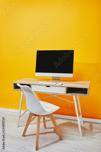 Computer with blank screen at wooden table and chair near yellow wall © LIGHTFIELD STUDIOS