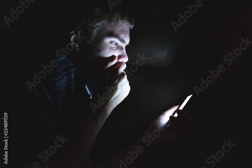 Teenager reading shocking news on his cell phone as he is lying on a couch in the dark.