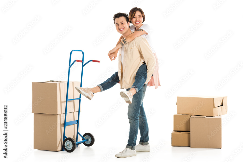 husband giving piggyback ride to wife near cardboard boxes and hand truck on background, moving to new house concept