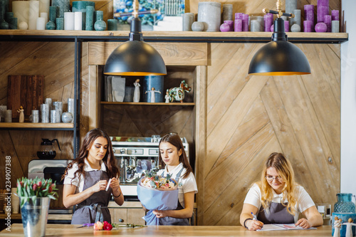 Team of three females florists working with flowers while standing behind wooden counter and creating beautiful bouquet  helping each other and writing in modern loft interior floral shop.