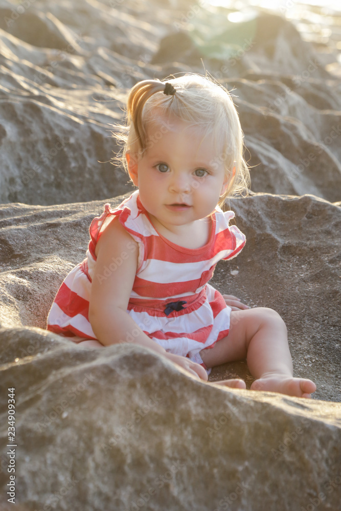 One-year-old baby girl in a red and white dress sitting on the rocky coast, spending time outdoors is important for little ones One-year-old baby girl in a red and white dress sitting on the rocky coast, spending time outdoors is important for little ones
