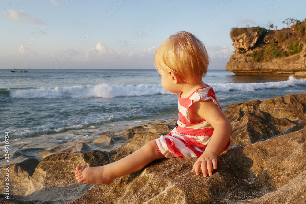 One-year-old baby girl in a red and white dress sitting on the rocky coast, spending time outdoors is important for little ones One-year-old baby girl in a red and white dress sitting on the rocky coast, spending time outdoors is important for little ones