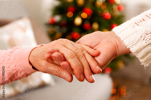 Elderly woman celebrating Christmas at home, with decorated holiday pine tree on background. Old lady at nursing home. Close up, copy space, cropped shot.