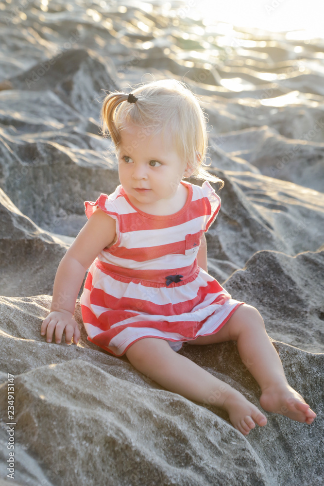 One-year-old baby girl in a red and white dress sitting on the rocky coast, spending time outdoors is important for little ones One-year-old baby girl in a red and white dress sitting on the rocky coast, spending time outdoors is important for little ones