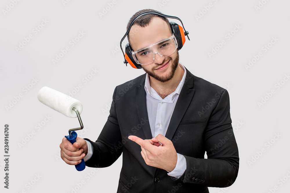 Confident young man in suit stands and points on paint roller he has in hand. Guy has headphones and glasses for protection. Usolated on white background. Confident young man in suit stands and points on paint roller he has in hand. Guy has headphones and glasses for protection. Usolated on white background.
