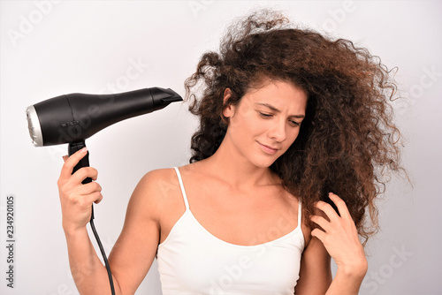 Cheerful lovely young woman in bathrobe standing and drying her curly hair with dryer over white background