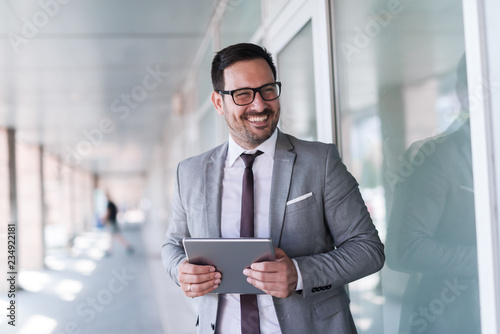 Close up of smiling businessman dressed in formal wear using tablet while standing near window outdoors. photo