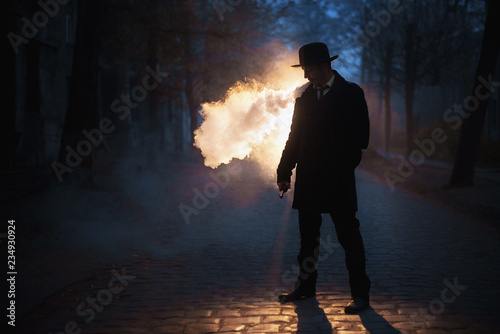 The man in hat smoke an electronic cigarette on the dark background outdoor. Vaponizer concept photo