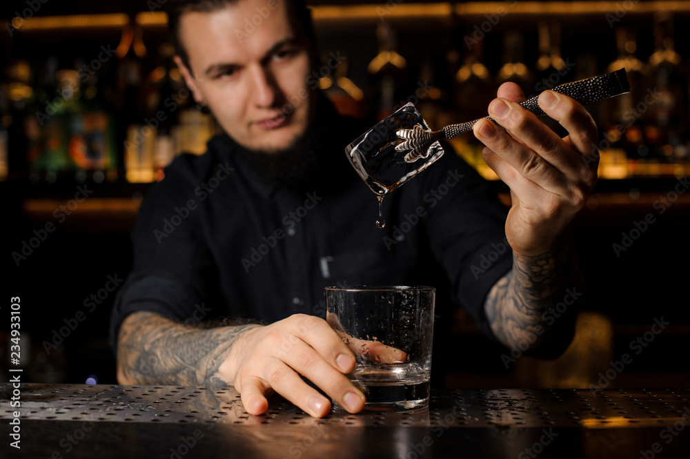 Young tattooed barman putting an ice cube into a cocktail glass