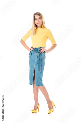 confident attractive woman in yellow shirt standing with hands akimbo and looking at camera isolated on white