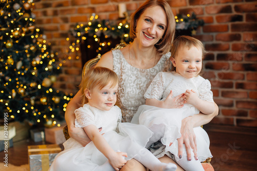 Mother with two cute little daughters in white dress sitting on the floor near christmas tree. Smiling woman playing with cheerful girls sisters. Happy new year